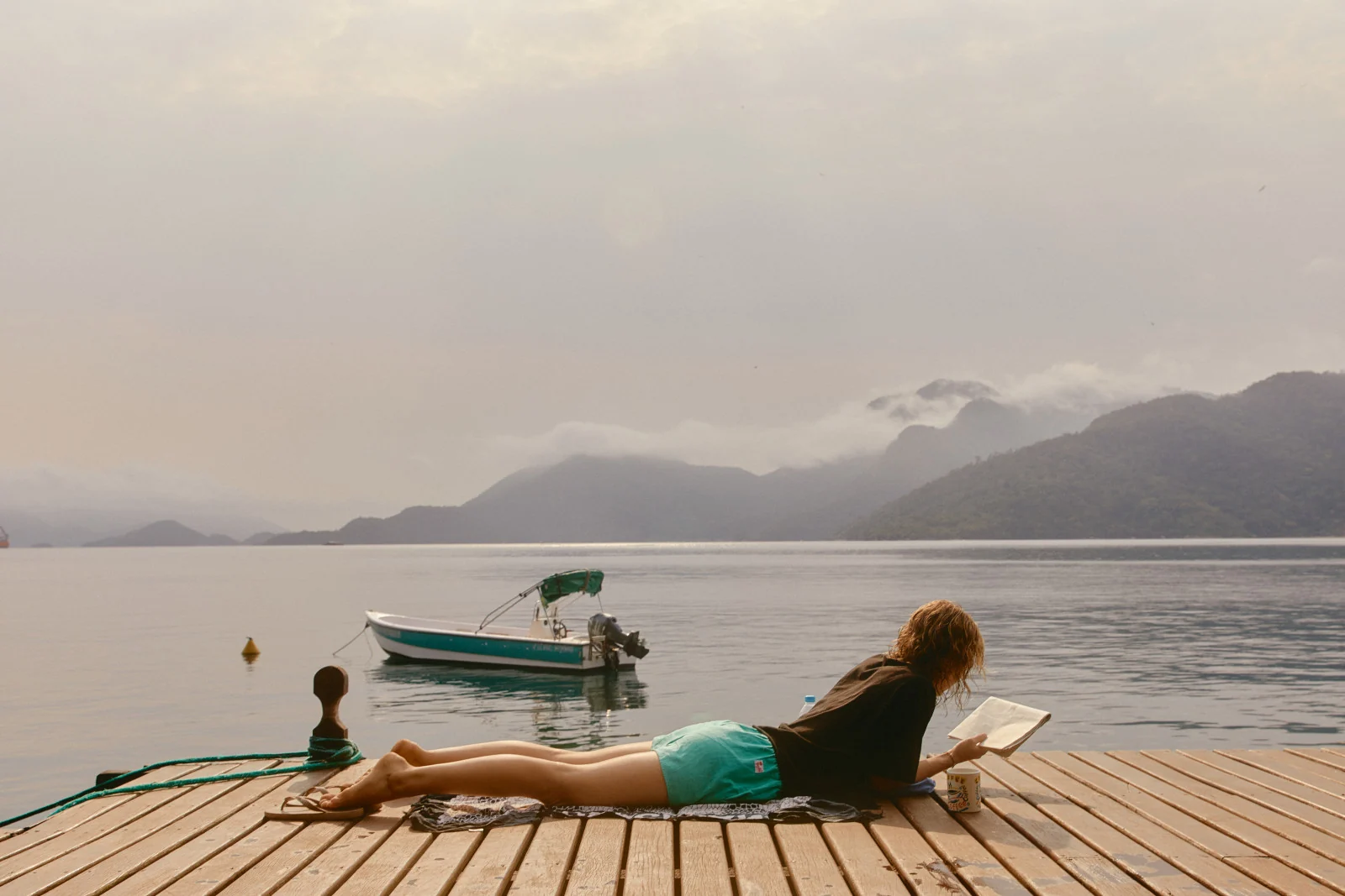 A woman reading on a wooden dock beside a still lake, a moored boat and mist-covered mountains beyond
